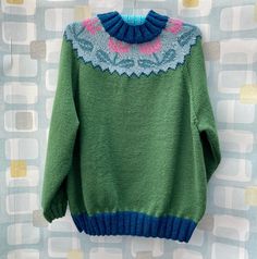 a green sweater with blue and pink details hanging on a wall next to a patterned background