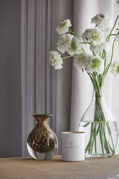 white flowers in a vase next to a candle on a table with curtains behind it