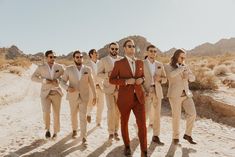 a group of men in tan suits and sunglasses walking across a desert road with mountains in the background