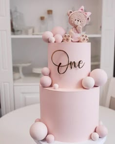 a pink cake with teddy bears on top and one written on the side, sitting on a white table