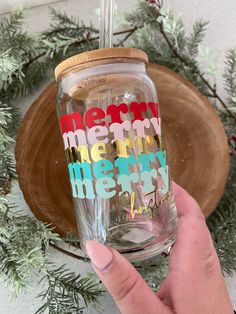 someone is holding up a glass with merry lettering on it and pine branches in the background