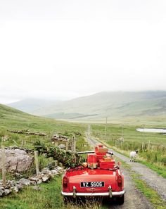 an old red car parked on the side of a dirt road next to a lush green field