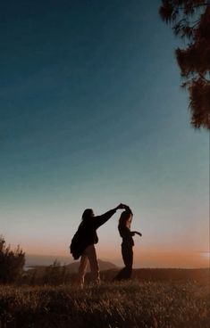 two people standing on top of a grass covered field next to a pine filled forest