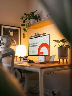 A well-lit home office featuring a minimalist white desk complemented by natural wood accents. The desk hosts a computer with a motivational message on the screen reading 'Don’t be busy. Be productive.' surrounded by lush potted plants and a stylish Batman poster. The space is designed for comfort and inspiration, with warm lighting and creative decor that encourages productivity. Desk Organization Tips, Minimal Workspace, Cozy Desk, Mac Studio, Architecture Model House, Cute Bedroom Decor, Studio Setup, Pc Setup, Contemporary House Design