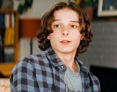 a young man with curly hair wearing a plaid shirt and looking at the camera while sitting on a couch