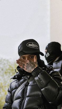 a man in a black jacket and hat covering his face with his hands while standing outside