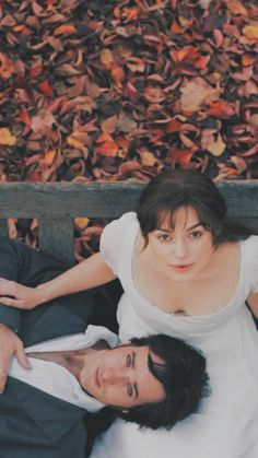 a man and woman laying on a bench in front of some fallen leaves with their arms around each other