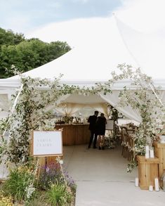 two people standing under a white tent with flowers and greenery on the side walk