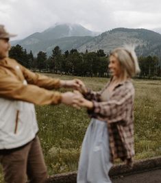 a man and woman holding hands while standing in front of a field with mountains behind them