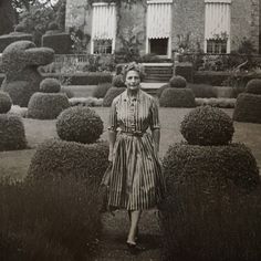 an old black and white photo of a woman standing in front of a house surrounded by bushes