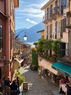 people sitting at tables on the side of a cobblestone street next to buildings