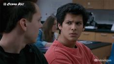 two young men sitting at a table talking to each other in an office setting with people working on computers behind them