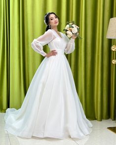 a woman in a white wedding dress standing next to a green curtain and holding a flower