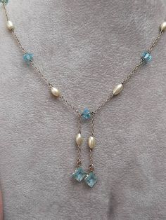 A 9ct gold  fresh water pearl, and blue crystal necklace. The necklace is not Hallmarked, but is stamped "375." 16 inch (406mm) chain, with two 1 inch (25mm) dangling pendants. The chain contains four crystals, and four grey shade fresh water pearls. It is joined at the bottom by a further blue crystal. Then suspended from the the centre in two strands is a further pearl, and a larger 5.5 x 5.5mm square cut blue crystal. The necklace is secured via a bolt ring clasp. Blue Pendant Necklace With Pearl Drop, Blue Crystal Necklace, Bolt Ring, Fresh Water Pearls, Fresh Water Pearl, Water Pearls, Square Cut, Blue Crystals, Crystal Necklace