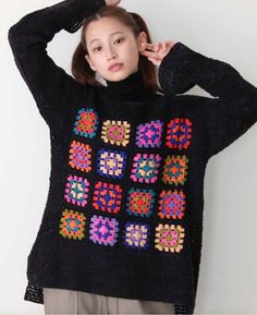 a woman wearing a black sweater with multicolored squares on it and holding her hands behind her head