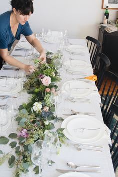 a woman setting a table with flowers and place settings on the long white tablecloth