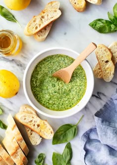 basil pesto in a white bowl surrounded by bread and lemons on a marble surface