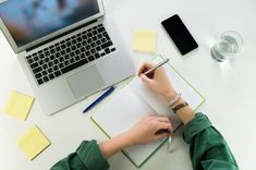 a person writing on a notepad next to a laptop computer and other office supplies