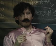 a man with a moustache holding a wine glass in front of a chalkboard