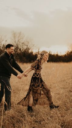 a man and woman holding hands while walking through tall grass in an open field at sunset