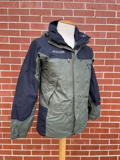 Image of a men's waterproof rain coat for sale. The jacket is made from a green and black waterproof material and was made by Columbia. It is a men's size small and is on a mannequin form with a white background for display. Sportswear Men, Waterproof Jacket, Zipper Pulls, Columbia Sportswear, Gore Tex, Front Zipper, Insulation, Columbia, Mens Jackets