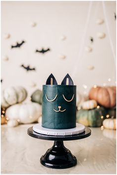 a green cake with a cat face on it sitting on a black stand in front of some pumpkins