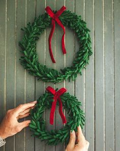 two hands are making a christmas wreath out of fake fir cones and red ribbon on a wooden surface