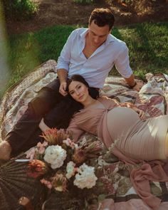 a man sitting next to a woman laying on top of a blanket in the grass