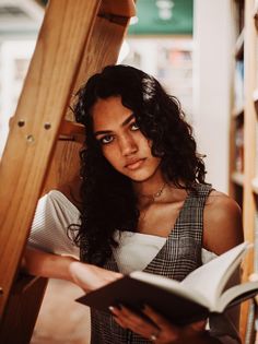 a woman reading a book while leaning against a wooden ladder in a library with bookshelves behind her