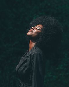 a woman with an afro standing in front of some trees and looking up at the sky