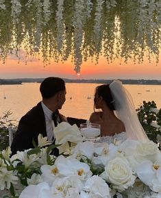 a bride and groom sitting at a table with flowers in front of the sun setting