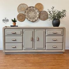 a gray dresser with baskets on top and plants in the corner next to it, against a white wall