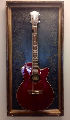 a red guitar hanging on the wall in front of a blue and black background with a gold frame