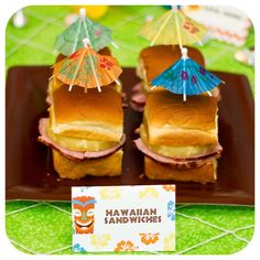 some sandwiches with umbrellas are on a brown plate and there is a sign that says hawaiian sandwhiches