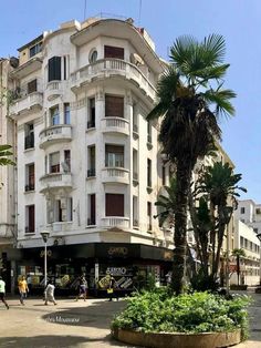 people are walking around in front of a white building with balconies and palm trees