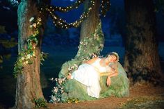 a man and woman are sitting on a bench in the woods with fairy lights strung around them