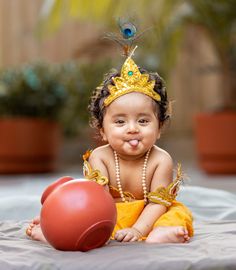 a baby sitting on the ground next to a red pot and wearing a gold crown