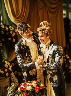 two men dressed in fancy clothing standing next to each other with flowers on the table