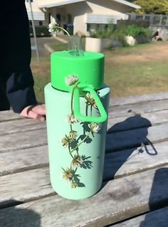 a green water bottle sitting on top of a wooden bench next to a hand holding a flower