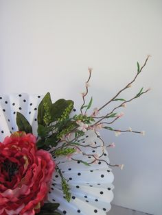 a white vase with red flowers and green leaves on the table next to a polka dot wall