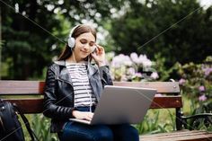 a woman sitting on a park bench using a laptop and listening to headphones