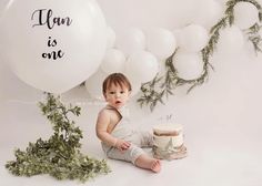 a baby sitting in front of a cake with balloons and greenery behind it that says, i am is one