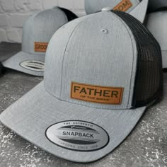 two hats sitting on top of a cement floor next to each other, one has a leather patch and the other has an orange sticker that says father