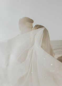 a man and woman standing next to each other near the ocean with veils over their heads