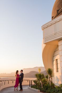 a man and woman standing next to each other in front of a building at sunset