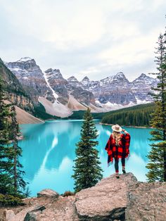 a woman standing on top of a rock next to a blue lake with snow covered mountains in the background