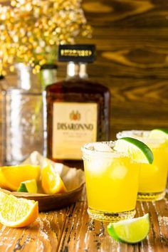 two glasses filled with lemonade and limes on a wooden table next to bottles