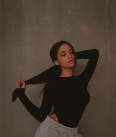 a woman with long hair standing in front of a gray wall wearing grey sweatpants and a black top