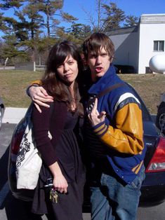 two people standing next to each other in front of a parked car with their arms around one another