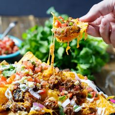 a hand holding a tortilla chip with meat and vegetables on it, over a bowl of lettuce